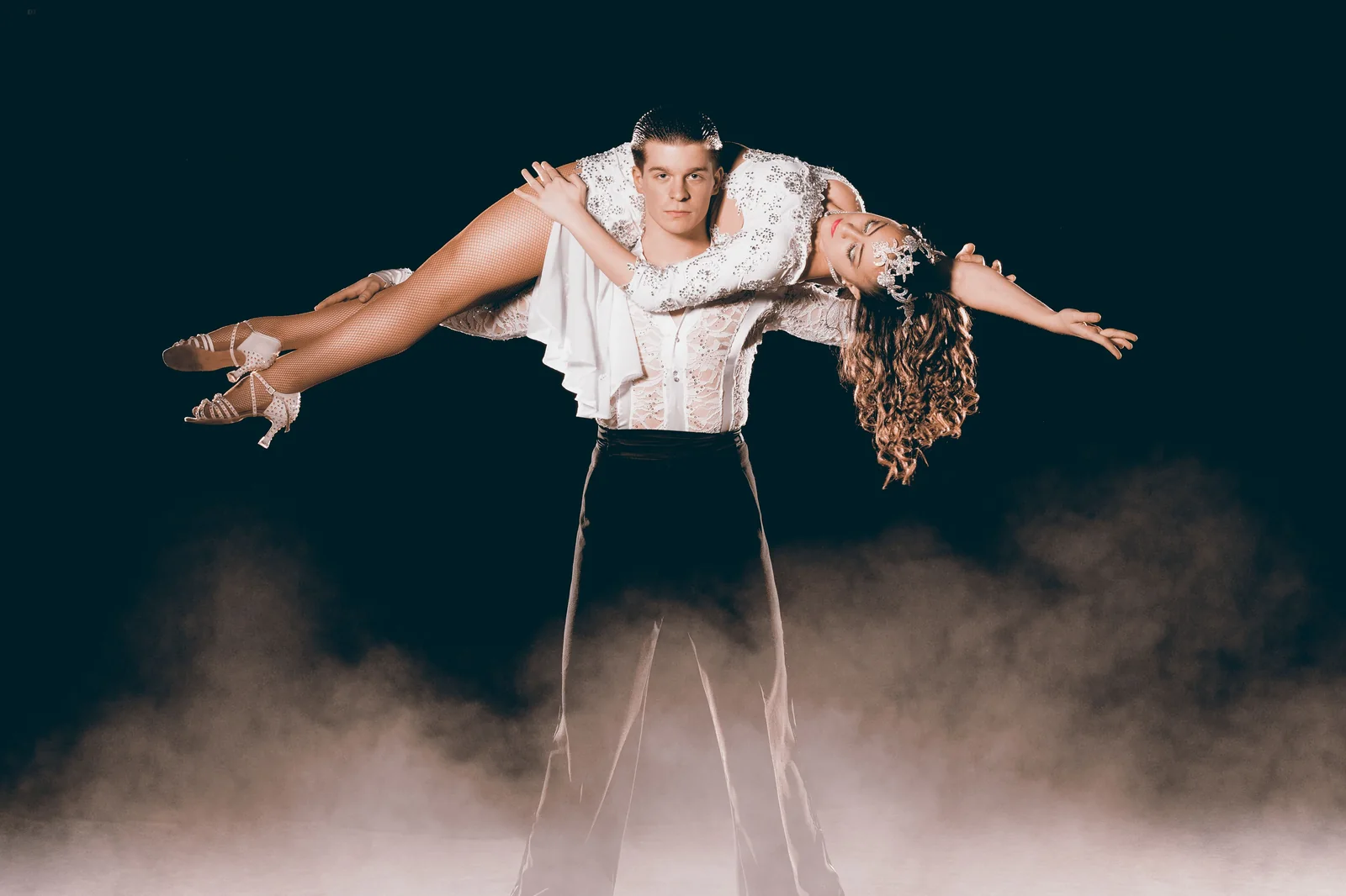 Dance partners performing an overhead lift on a dark stage with theatrical fog