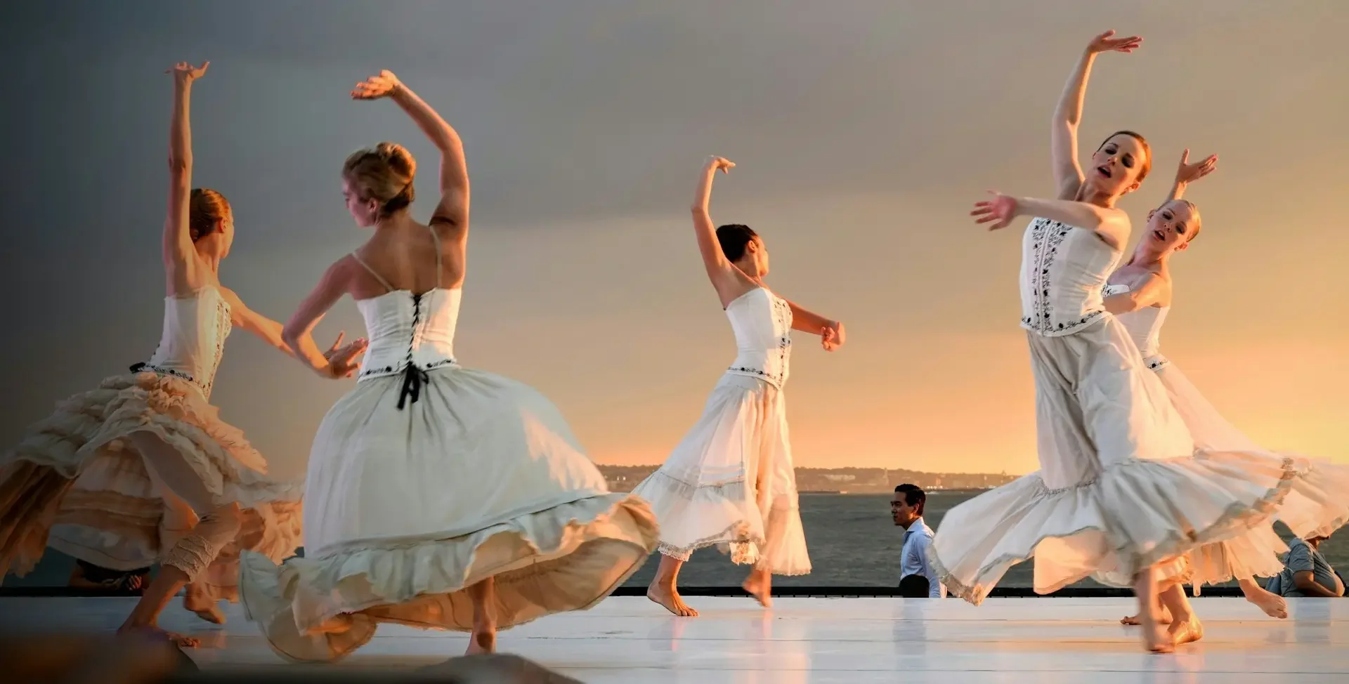 Dancers in white costumes performing on an outdoor stage at sunset by the water