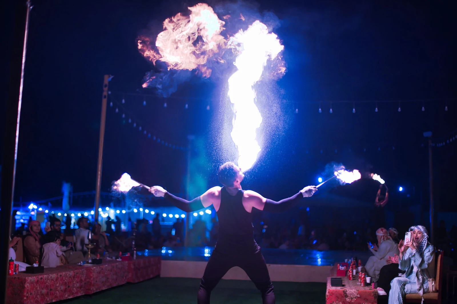 Fire performer breathing a plume of flame at an outdoor night show with seated guests