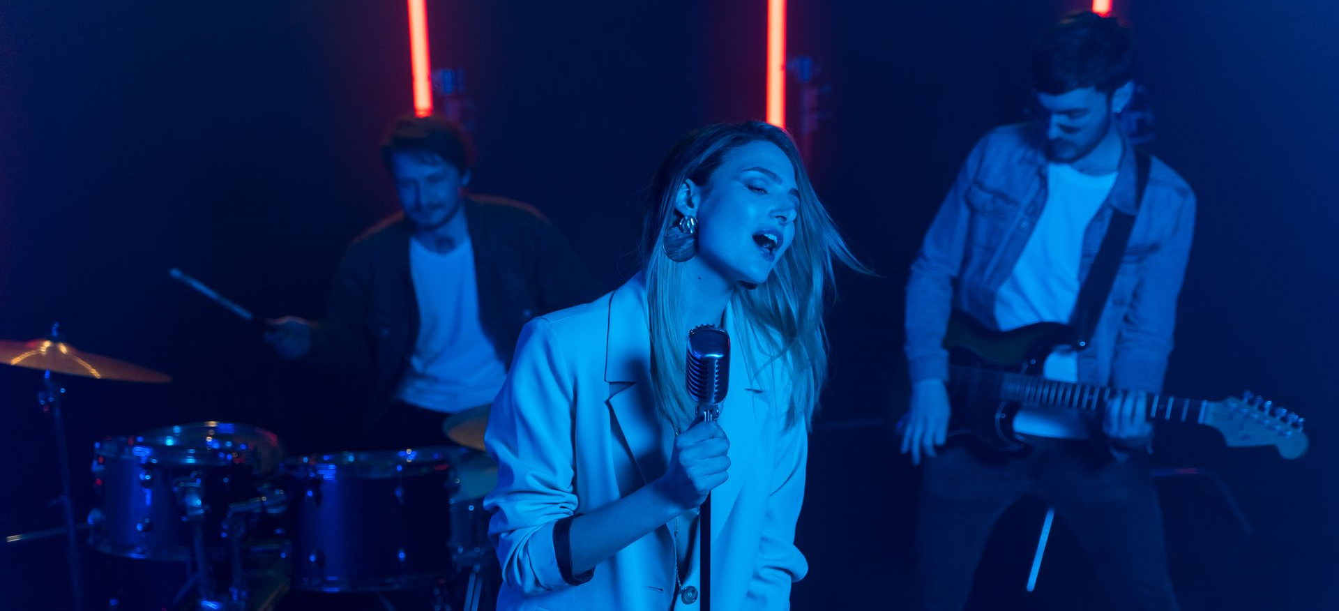 Live band performing on stage with vocalist, drums, and guitar under blue and red lights